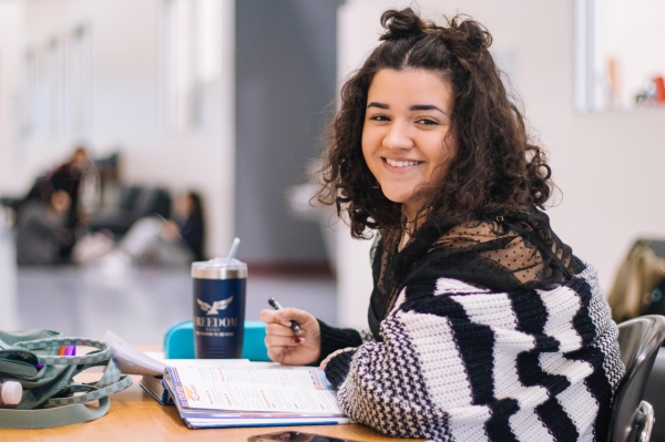 A girl smiling. She&#039;s sat at a desk with an open book on top of the desk.